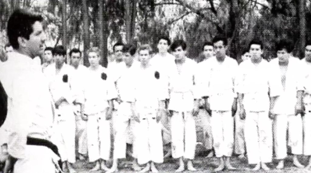 Foto en blanco y negro de un grupo de jóvenes karatekas formados frente a su sensei en medio de un bosque